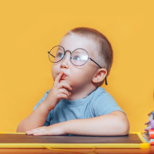 A young child wearing glasses sits at a desk with a finger on their lips, surrounded by colored pencils, apples, and stacked books, against an orange background.
