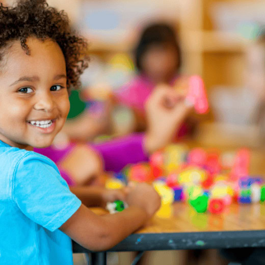 A young child in a blue shirt smiles at the camera while playing with colorful building blocks at a table in a classroom.